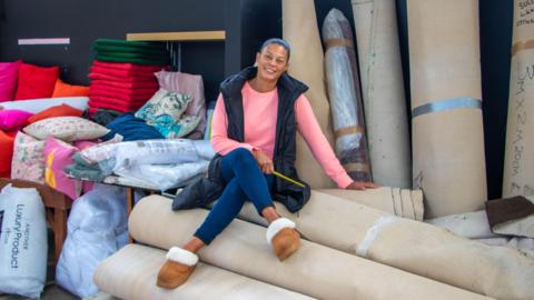 A woman, Pia Honey, is sat on a pile of rolled up carpets in front of a pile of other carpets and cushions and she is smiling at the camera. Pia is wearing a pink top, black gilet, jeans and brown boots.