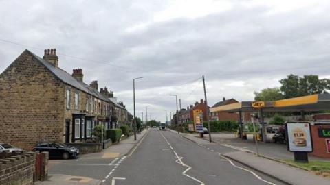 A long, straight suburban road, with terraced houses on one side and a petrol station on the other. 