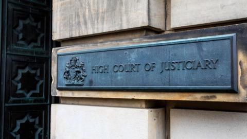 A grey metal sign on a sandstone exterior wall. The sign features a royal crest and the words: "High Court of Justiciary".