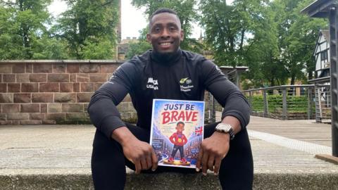A black man in black running gear holding a children's book with the title Just Be Brave