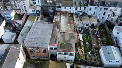 A drone photo of the former nightclub building that is surrounded by three and four storey buildings. The flat roof of the former club is flat and there are hedges in the courtyard area.