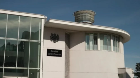 Image of Exeter Crown Court. It is a close up of a white building which has large glass panels on the left-hand side. A large black plaque says 'Exeter Law Courts'