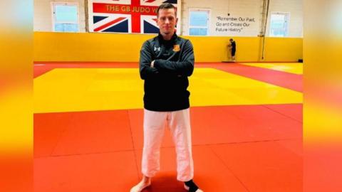 A man with short dark hair, wearing a dark jumper and white trousers, stands with his arms folded on an orange coloured mat in a large hall. A yellow mat can be seen behind him and a Union Flag is hanging on the wall.