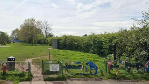 A park area off Westfaling Street in Hereford. There are barriers and children's play signs and there is a waste bin. There are hedges around an area of open green space.
