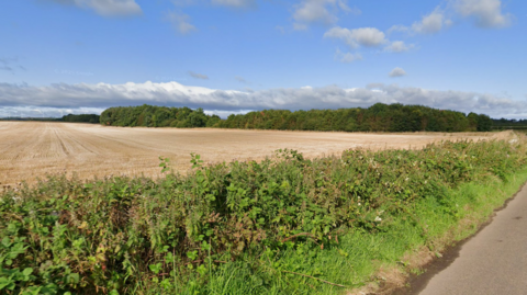 Google Streetview of woodland, seen from the road. Close up are a verge and a shrubby hedge with a small section of rural road. Beyond those is a recently harvested crop field with a small woodland beyond. The sky is blue with a few white clouds and it's a sunny day.