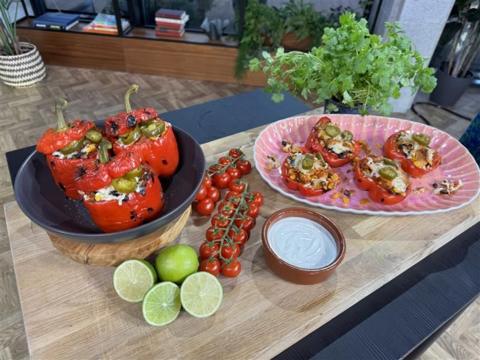 Stuffed red peppers in black bowl with green jalapenos sticking out of the top to the left. There is three half limes and one full lime in front of the black bowl. Two vines of red cherry tomatoes behind the limes to the side of the bowl. Centre left there is a small bowl of sour cream. Behind the sour cream to the right there is a pink oblong plate with stuffed pepper halves with a pot of coriander behind it to the right. All of these items are on a wooden board.