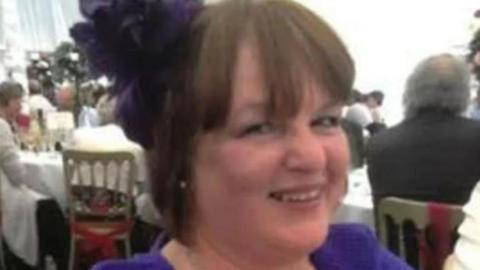 Linda Stockdale pictured at what appears to be a wedding, wearing a purple outfit and a purple hat while smiling at the camera