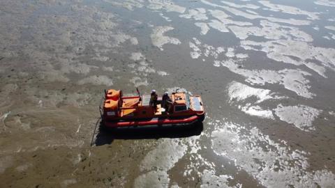 An orange hovercraft glides across wet sand with some areas completely covered in water amid a rising tide. There are two people on the hovercraft.