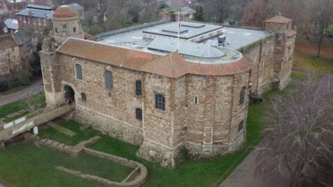 An aerial view of Colchester Castle, taken from a drone. The castle is made of yellow stone walls with a mostly flat roof. There is grass surrounding the castle with exposed foundations, and there is a drawbridge going into the main entrance of the castle.