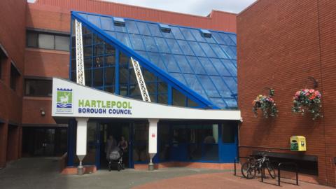 The council building is a blue glass design surrounded by a red-brick building. A white and green sign above the automatic sliding doors reads: "Hartlepool Borough Council".