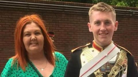 Jack standing in his uniform with his mum on his left hand side. Jack is wearing a full Household Cavalry uniform, including a metal chest plate with gold chains and a white sash across it, and a back jacket. He has short blond hair. Laura has dyed red straight hair and is wearing a green dress with black polka dots. 