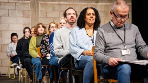 Actors sat in a line with one or two people in each line, as if they are on an aeroplane. The man in the front is wearing a lanyard and looking at a script. Most people are wearing jeans and they have a range of different tops on. 