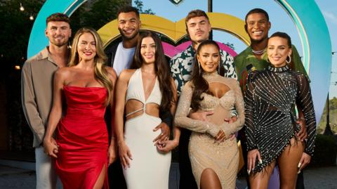 The eight Love Island finalists stood smiling in front of a blue, pink and yellow heart. 