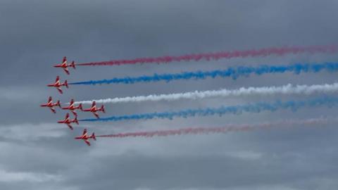 The Red Arrows in formation with red, white and blue smoke trailing behind the red planes.