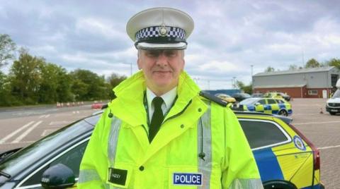 Jack Hadley is smiling into the camera. He is wearing a neon yellow/green high vis jacket and it says POLICE. He is also wearing a police hat which is white. There is a car behind him, which is a blue and yellow police car. 