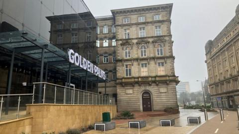 A quiet street lined with both modern and historic buildings. On the left, a sleek glass structure with a sign reading “GOLDEN PALMS” contrasts with the ornate stone building beside it, which features tall windows and decorative architectural details. The pavement looks wet, and the sky is overcast.