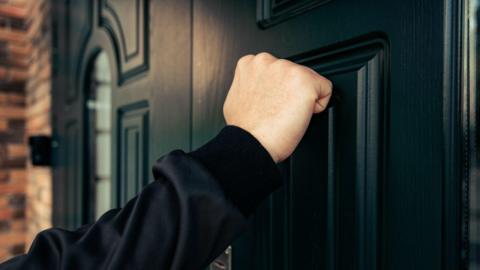 A hand knocking on a black door. The person is wearing a black long sleeve jumper.