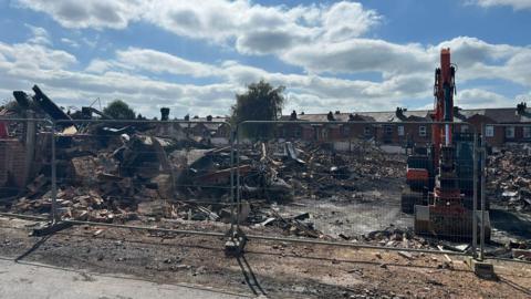 A site of a demolished building behind metal fencing. Huge piles of rubble are scattered across the site and on the right, a yellow digger is parked. Behind the site is a row of terraced houses.