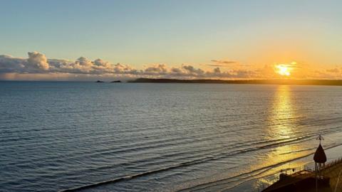 The sun setting over the sea on the right hand side with a row of white clouds across the horizon