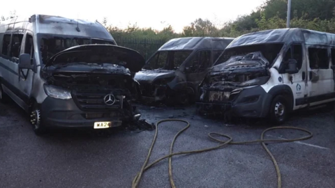 Three minibuses in a compound are completely blackened and burned out. A yellow fire service hose is on the ground near them.