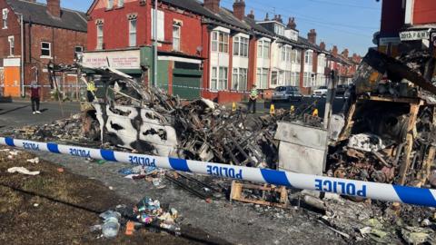 A burnt-out bus sits behind some police tape 