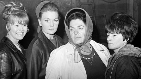 A black and white photo of four women, two of them wearing headscarves. The woman on the left is smiling and the other three have serious expressions on their faces.