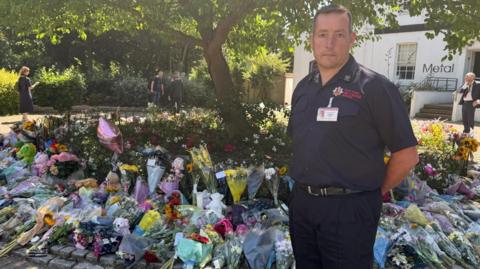 A man standing in his uniform with a background of flowers spread across a tree. Teddy bears are also being left. The man has a sombre expression on his face and he has his hands behind his back.