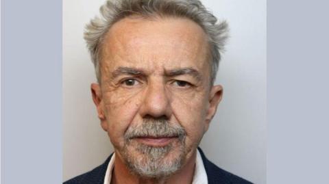 A custody mugshot of Aleksandr Franczyk, who has grey hair and a goatee, and is wearing a blue blazer and a white shirt