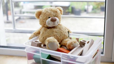 A teddy bear is sat on top of a plastic box of wooden blocks. 