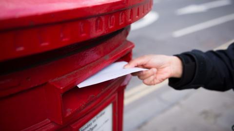 A hand is seen posting a letter in a red post box