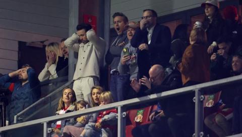 Rob and Ryan at the Wrexham stadium looking disappointed as the team lost against Chelsea. There are other fans in the stands as well.