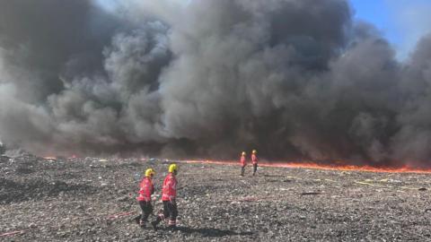 Four firefighters are wearing yellow helmets, red jackets and black trousers. They are standing on a landfill site which is on fire. There is a huge black cloud of smoke.