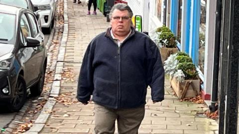 A man with greyish black hair slicked back and glasses in a blue zip up top and beige trousers walks along a cobbled street with shops to one side