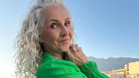 A woman with curly silver hair, wearing a green dress, turns to face the camera and smiles