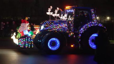 A blue tractor has Christmas lights attached to it and at the front has an inflatable Christmas tree and Santa.