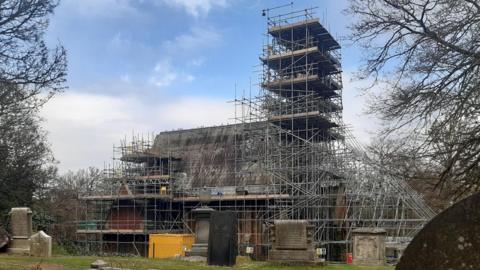 The chapel which is covered in scaffolding. It has a tall spire which is also covered. There are about six, large gravestones dotted on the green field in front of the chapel.