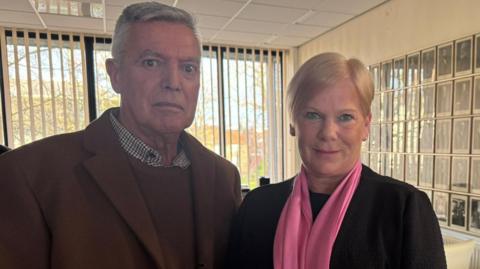 Rachael O'Brien and her husband Joe. Mrs O'Brien is smiling into the camera and has short, blonde hair and is wearing a black top with a bright pink scarf around her neck. Her husband is standing to the left and has short, grey hair and is wearing a brown coat, shirt and a checked shirt. They are standing in a large room with has a wall covered in black and white portraits of people. There is a large window with vertical blinds behind them.