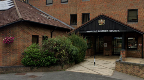 A large brown building with shrubs and plants on the side. 