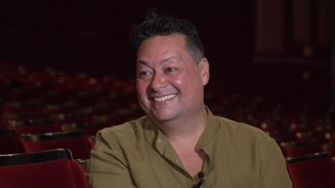A man with short black hair smiles while sitting in theatre stalls. He is wearing a green shirt