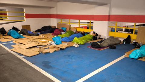A group of people lying in sleeping bags on top of cardboard sleeping rough in a car park for charity.