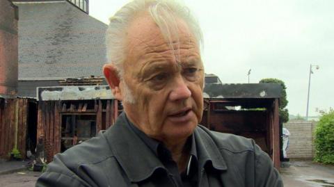 Fr Paddy O'Kane, wearing a black jacket, and with silver hair and long sideburns, stands in front of a church badly damaged in a fire in 2019