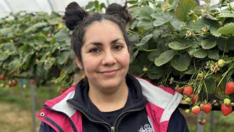 A woman with dark hair tied up in two buns above her head with a pink jacket in front of a row of plants with some strawberries on