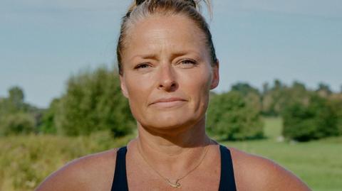 A head and shoulders photo of Laura Reineke, a woman with blonde hair tied in a bun. She's wearing a black swimming costume and a necklace with a mermaid on. She's looking straight at the camera with a half-smile, with trees and a blue sky behind her.