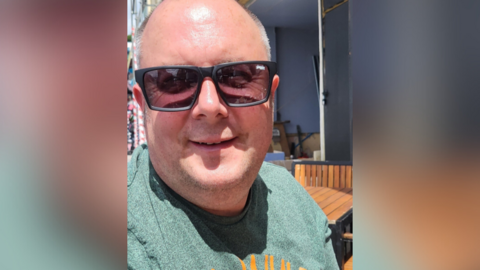 A man sits in the sun at an outside table. He is wearing a green t-shirt and sunglasses and smiling. 