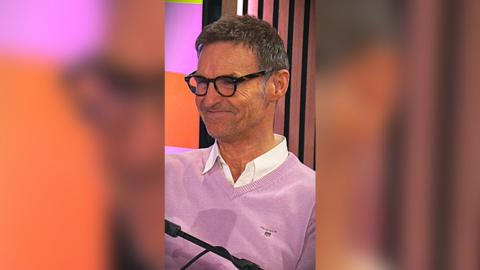 Marti Pellow in a light pink v-neck knit jumper with white shirt collar poking out the top. He is wearing dark framed glasses and is smiling.