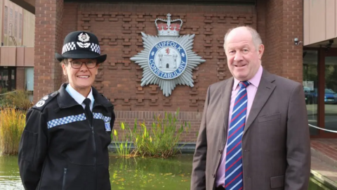 Rachel Kearton in police uniform next to Tim Passmore, who is wearing a grey or brown suit over a pink shirt and striped tie. They are standing in front of Suffolk Police's headquarters. The force logo can be seen behind them on a brick wall, in front of an ornamental pond.