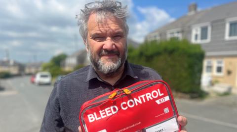 A man with grey hair and a beard looking at the camera, he is wearing a dark grey shirt and has glasses pushed back on his head. There is a street with houses in the background and he is holding a red case which says bleed control kit in white lettering