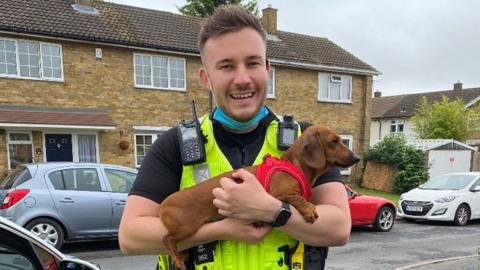 A police officer, young, is wearing a uniform and holding a puppy next to a police car on a residential street