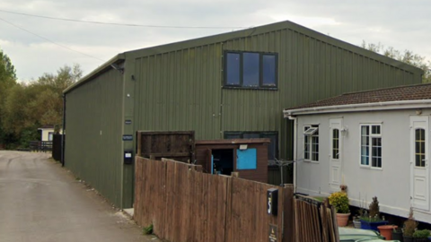 A large green barn with one window at the top. In front there's a white mobile home and a wooden fence. They are both on a lane, with some trees in the distance. 