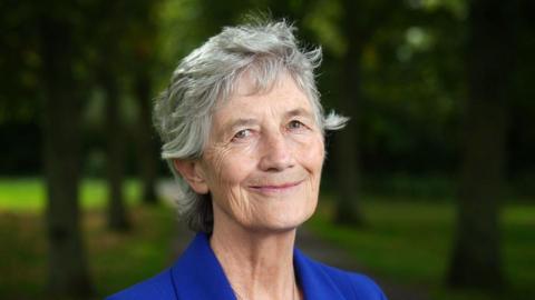 Catherine Connolly has grey hair down to the bottom of her neck and is wearing a blue suit jacket. She is standing outside with a number of trees in the background.